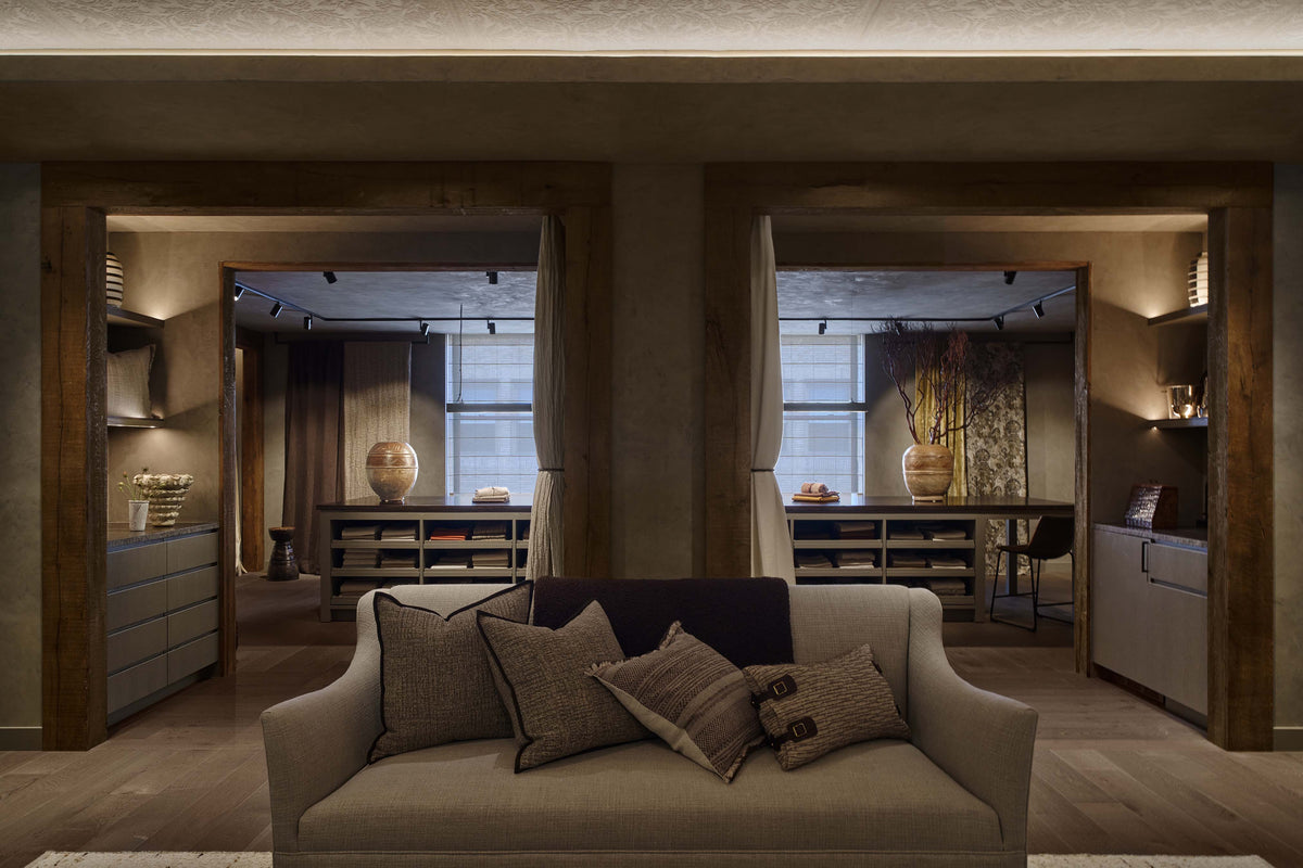 A modern beige fabric sofa with multiple textured throw pillows, positioned in a warm, contemporary interior with rustic wood trim and open archways leading to other rooms.