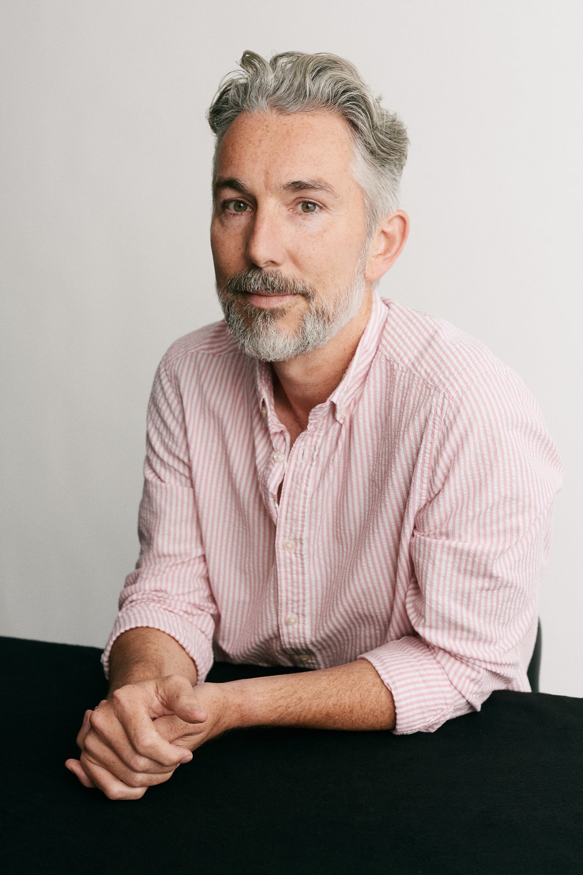 A man with gray hair and a beard wearing a pink and white striped button-down shirt, sitting with his hands clasped on a black surface against a light gray background.