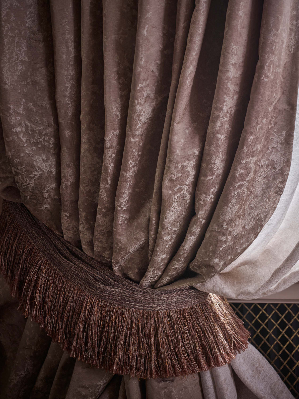 Close-up of a draped curtain in a textured taupe-brown fabric with a matching fringed tieback, next to a sheer off-white inner curtain.