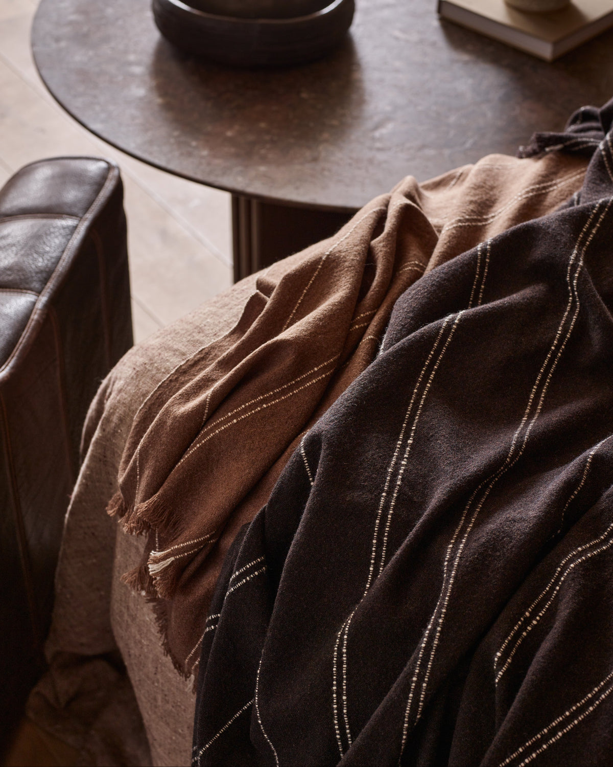 A close-up of draped textiles in a styled interior setting, featuring a dark brown throw blanket with thin light vertical stripes and a tan woven throw draped over a chair beside a round dark table.