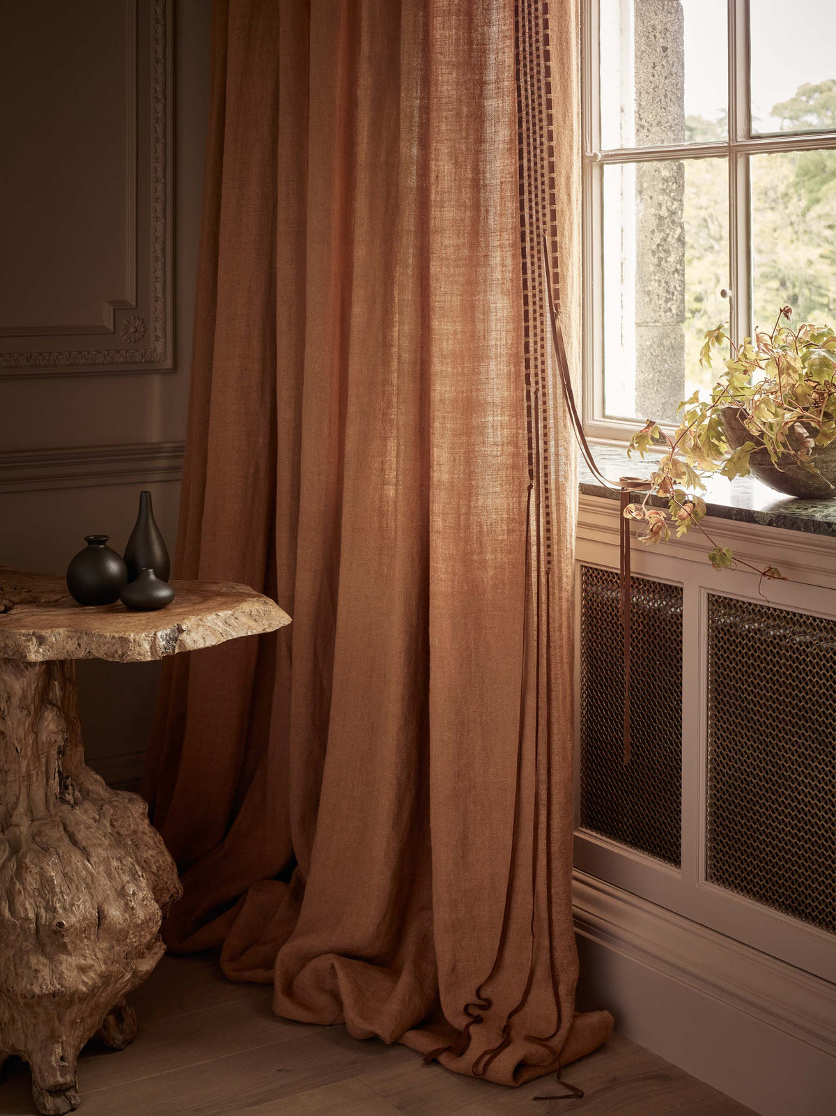 A pair of floor-length rust-colored linen curtains hanging by a window in a styled room with a rustic wooden side table and a potted plant on the sill.
