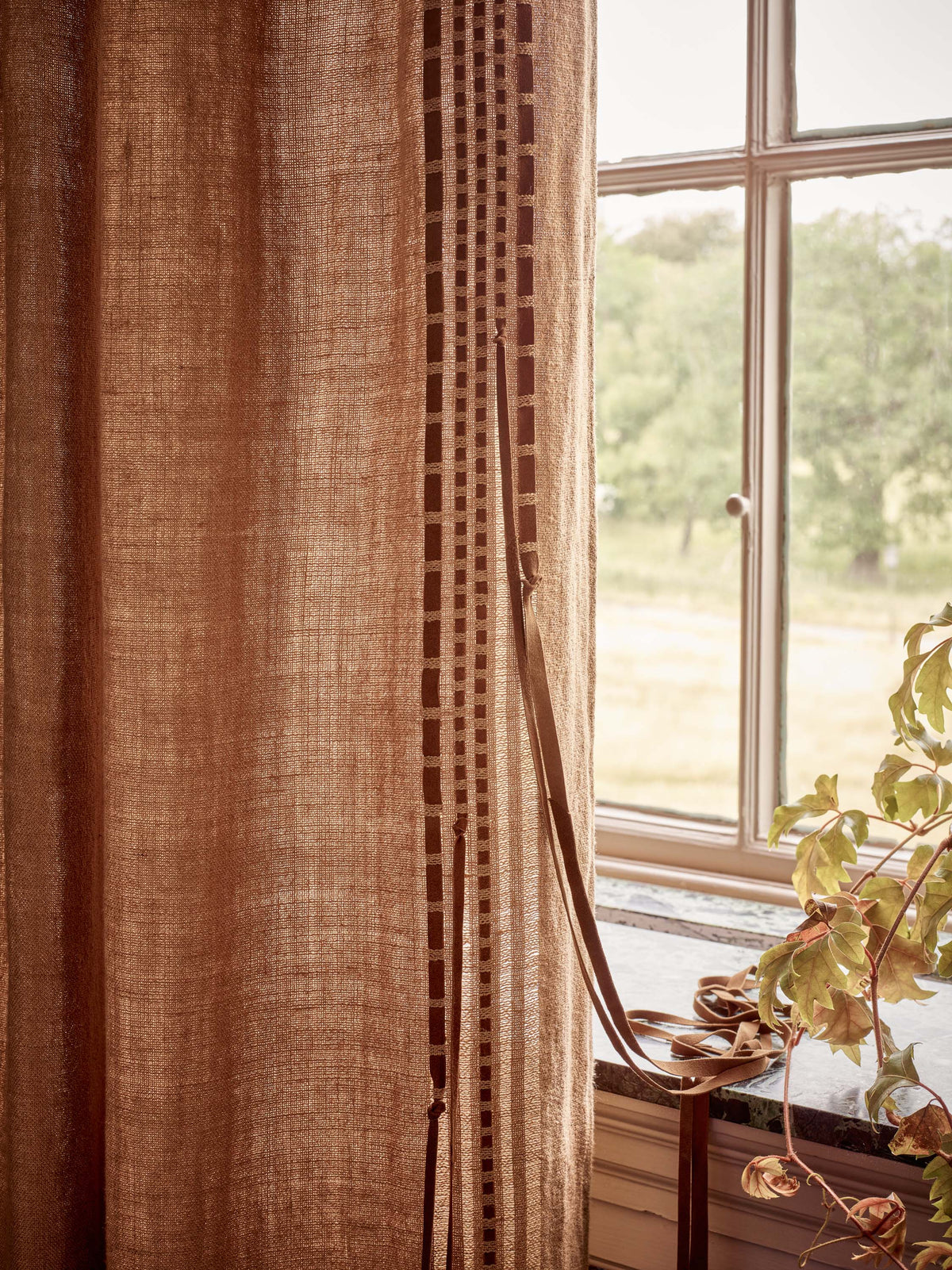 A close-up of a textured brown curtain panel hanging next to a window, with a decorative vertical stripe detail and a plant visible outside.