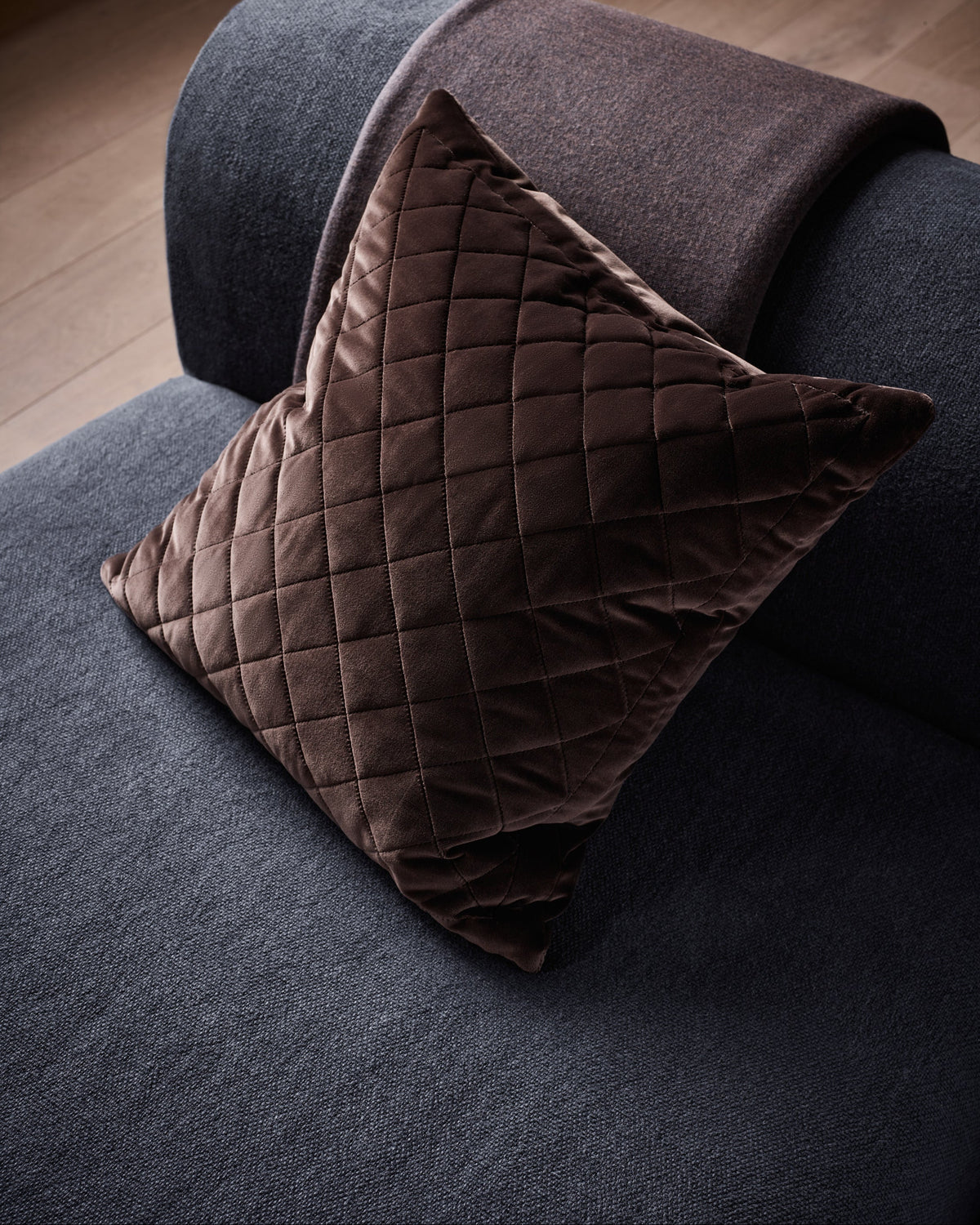 A dark brown quilted velvet throw pillow resting on a dark gray sofa with a matching brown throw blanket draped over the arm, set against a wooden floor background.