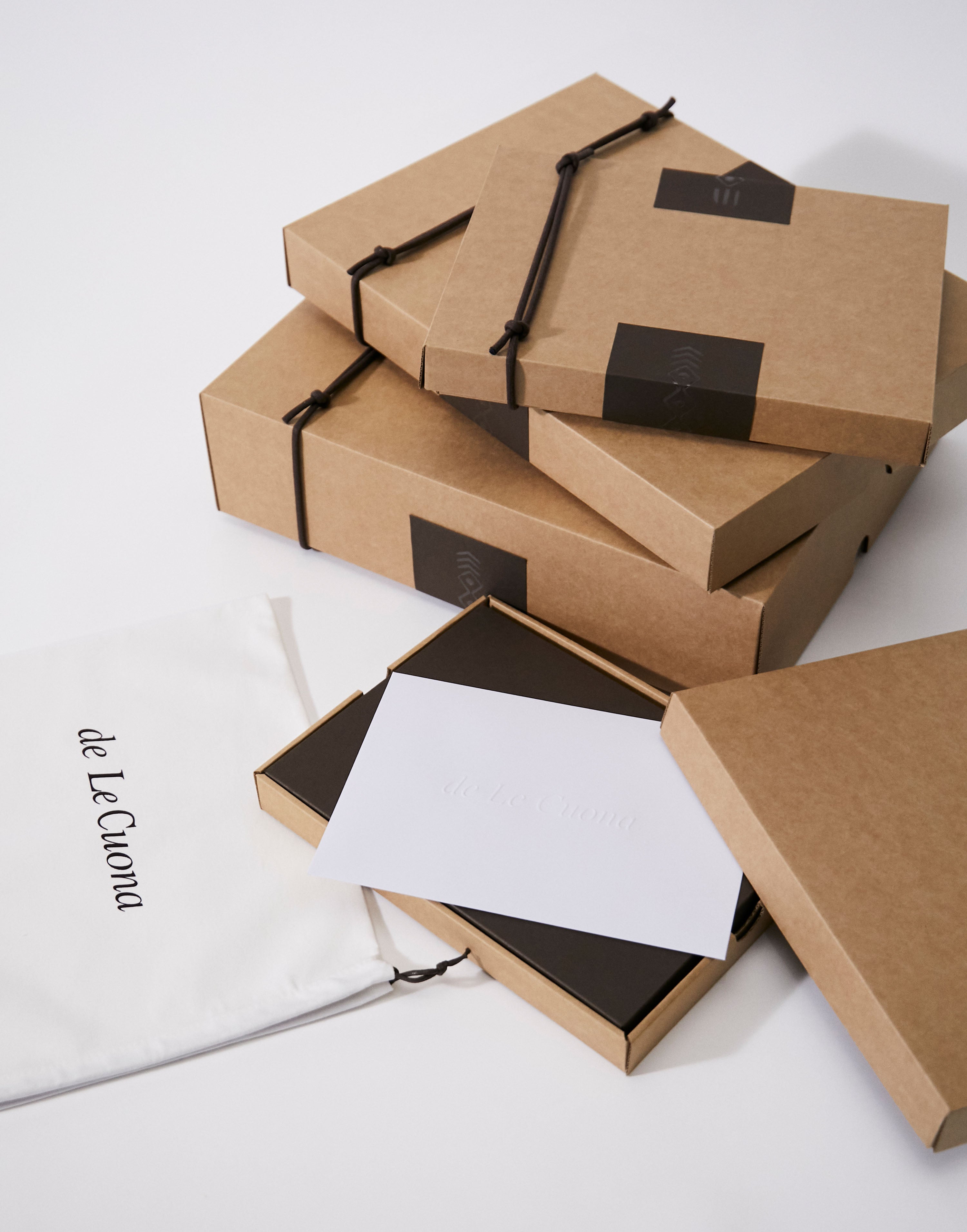 Stack of brown cardboard boxes with black ribbons on a white background, featuring the brand 'de Le Cuona'.