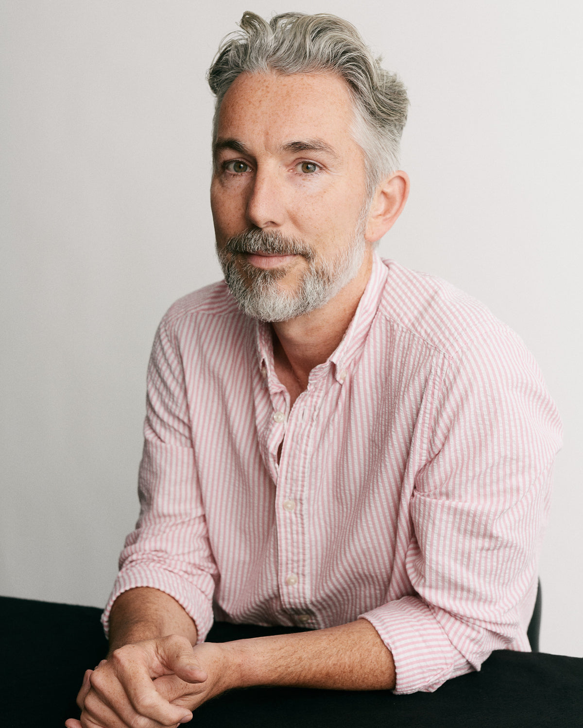 A man with gray hair and a beard wearing a pink and white striped button-down shirt, sitting with his hands clasped on a black surface against a light gray background.