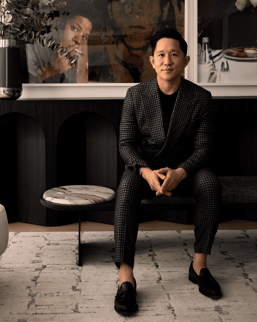 A man wearing a black and white checkered blazer and matching trousers sits on a dark bench in a modern interior setting with framed art and a patterned rug.