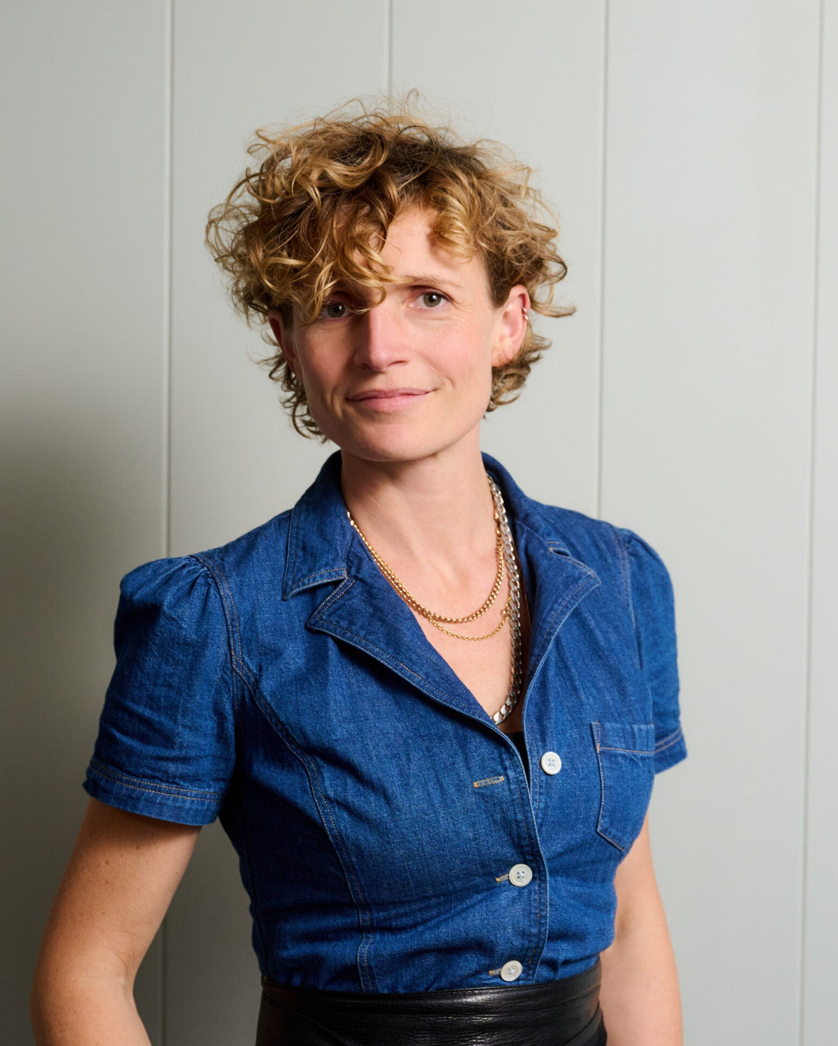 A woman with short curly blonde hair wearing a blue denim short-sleeve button-up shirt and a black leather belt, standing against a light gray paneled wall.